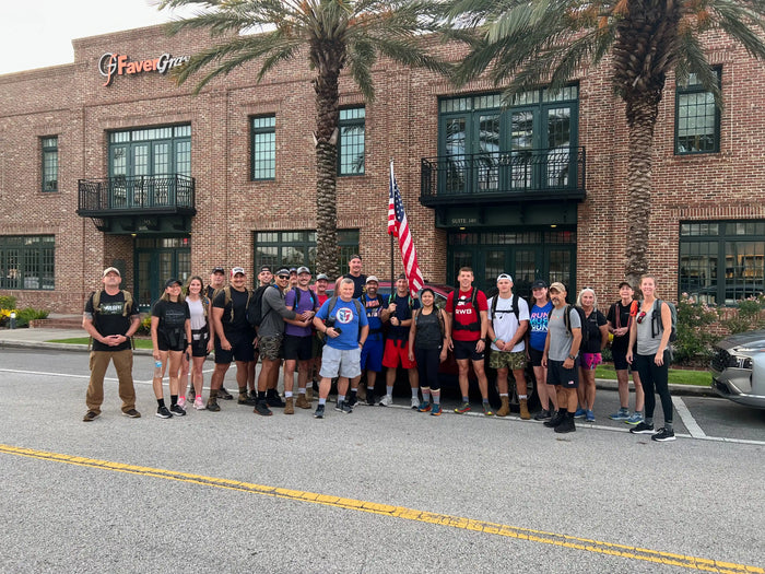 Group of GORUCK participants with rucksacks and American flag outside brick building, palm trees