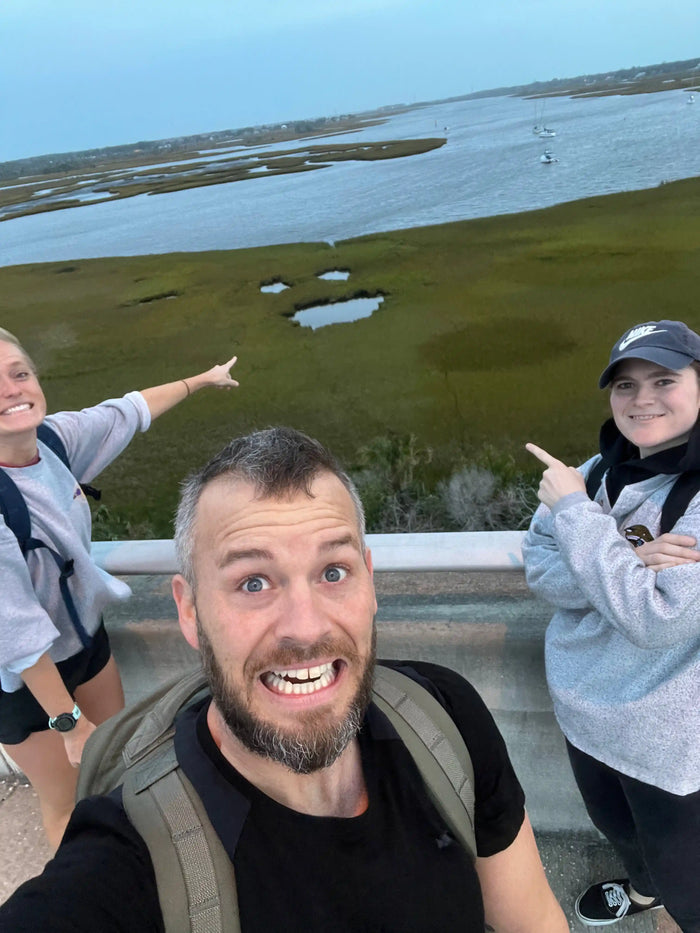 Three people with rucksacks smiling on bridge, pointing at marshland with GORUCK gear in view