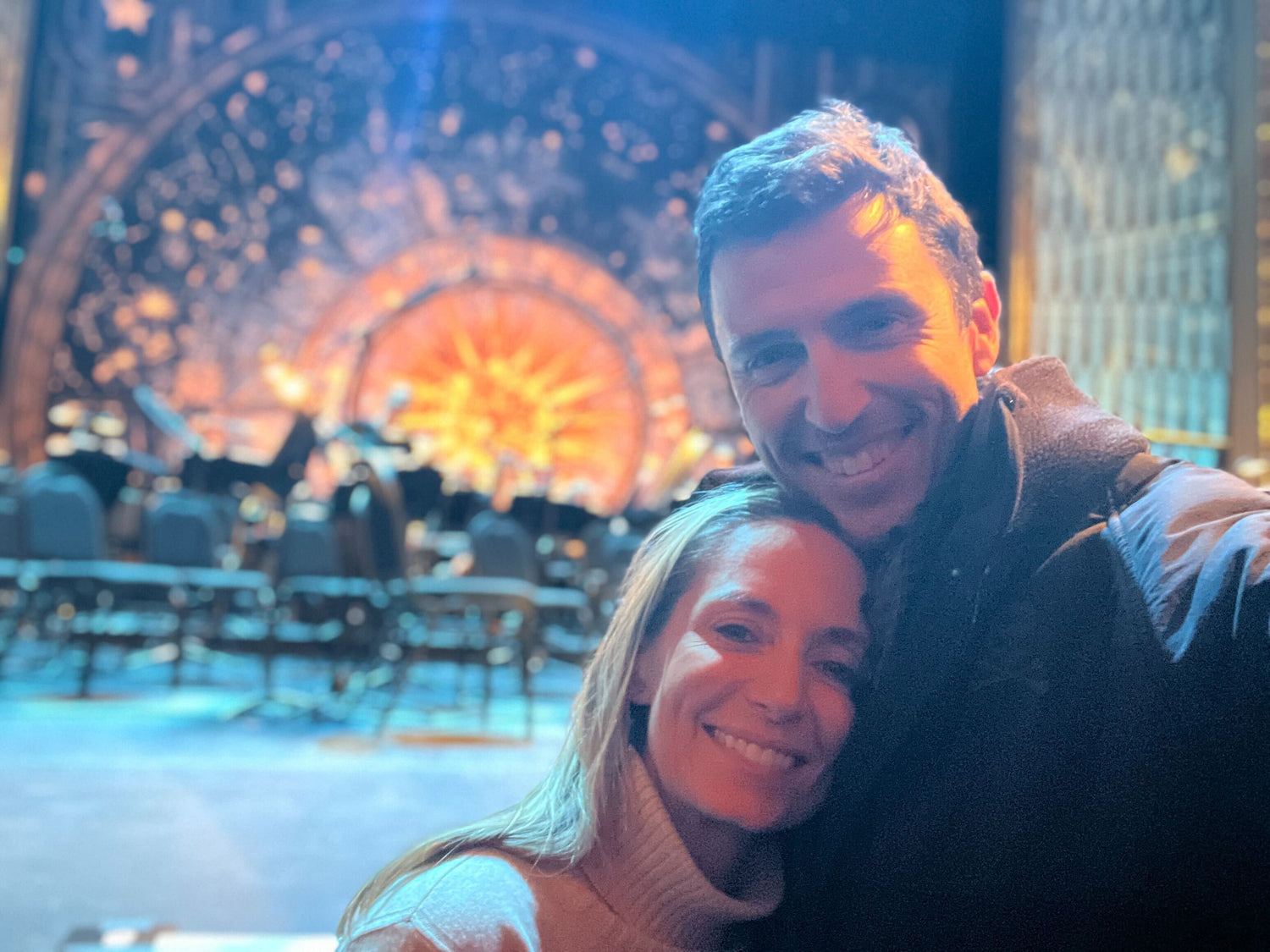 Smiling couple taking a selfie in front of illuminated stage with empty chairs and starry backdrop.