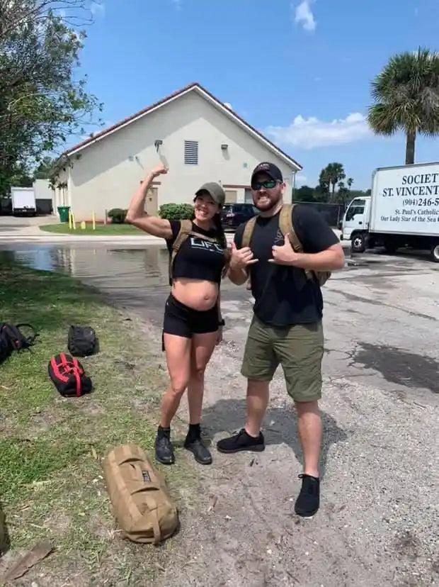 Two people with rucksacks posing outdoors by gear bags, promoting GORUCK rucking event.