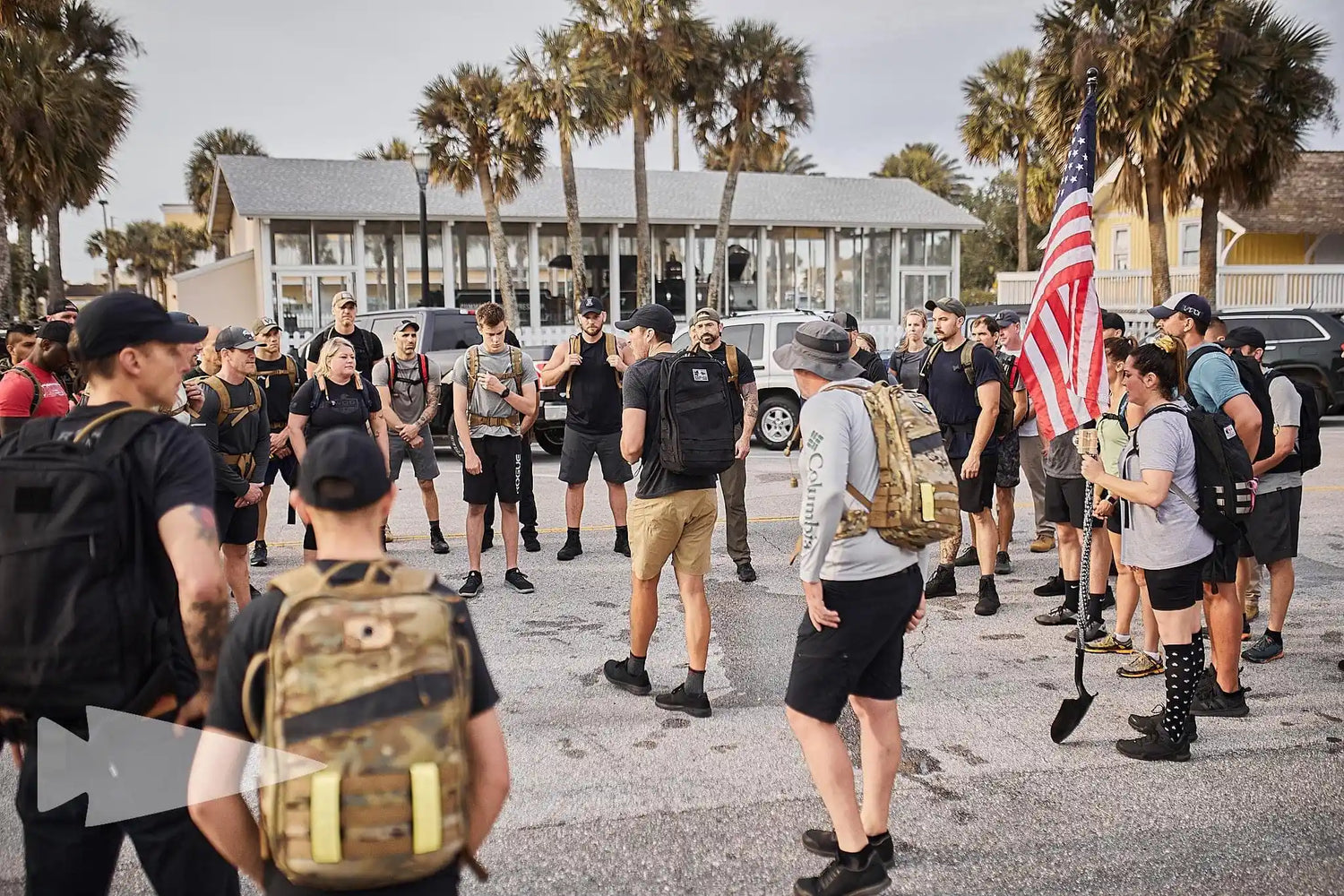 GORUCK rucking group gathering outdoors, wearing backpacks, American flag, palm trees in background