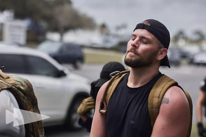 Man rucking outdoors with GORUCK backpack, wearing black tank top and cap, focused expression