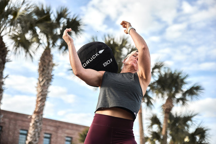Woman lifts GORUCK 80lb sandbag outdoors, palm trees in background, fitness gear focus