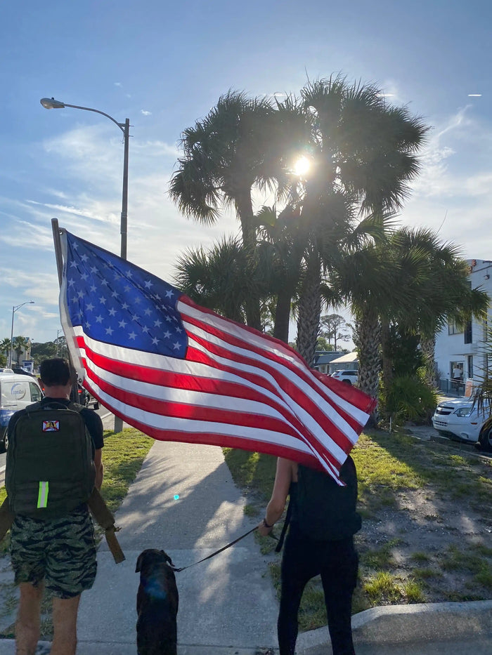 GORUCK rucking event participants carrying USA flag and dog, walking on palm-lined street