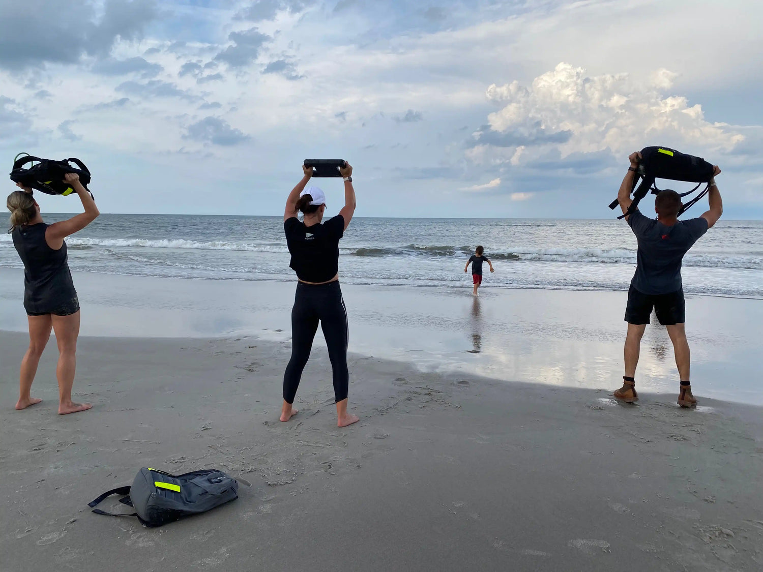 Three people rucking with GORUCK backpacks on a sandy beach by the ocean under cloudy skies.