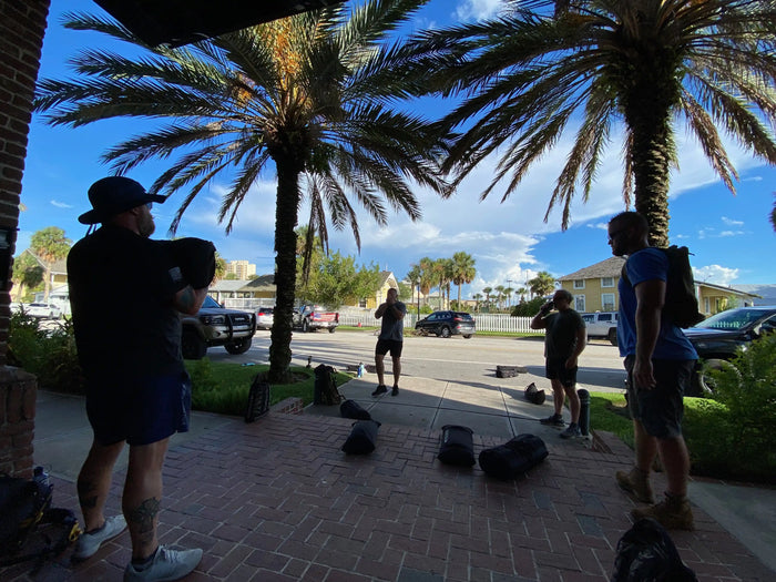 Group training with GORUCK backpacks outdoors under palm trees, sunny day, urban setting
