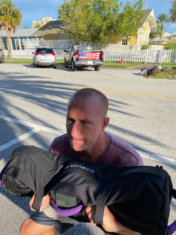 Man outdoors lifting GORUCK rucksack sandbag in bright urban setting with parked cars