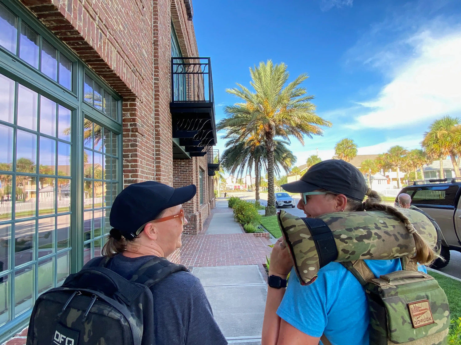 Two people rucking with GORUCK gear and backpacks on a sunny palm-lined sidewalk.