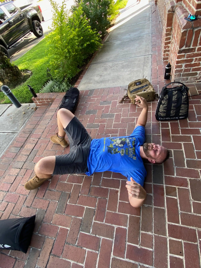 Man in blue t-shirt resting on brick walkway with GORUCK backpack and gear outdoors
