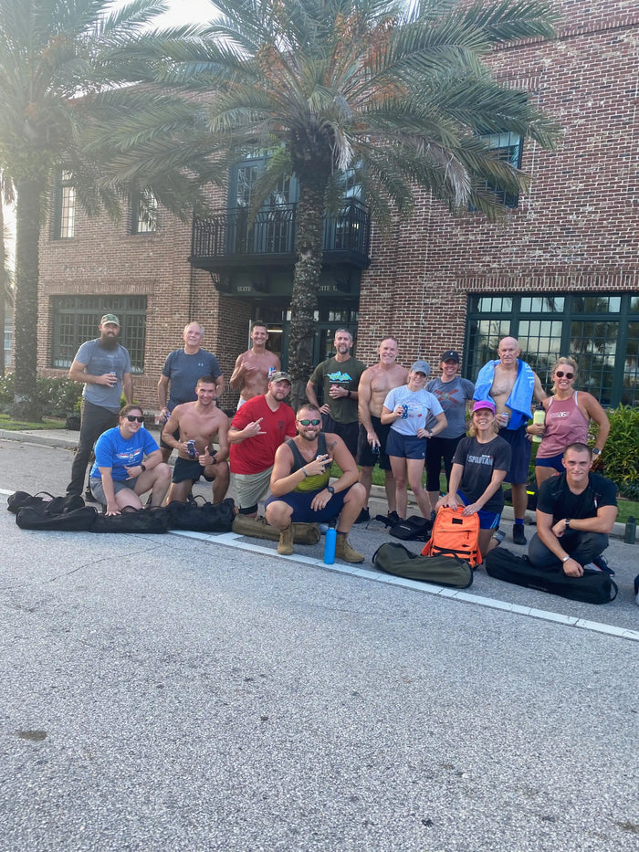 GORUCK team group photo outdoors with rucksacks, palm trees, and brick building background
