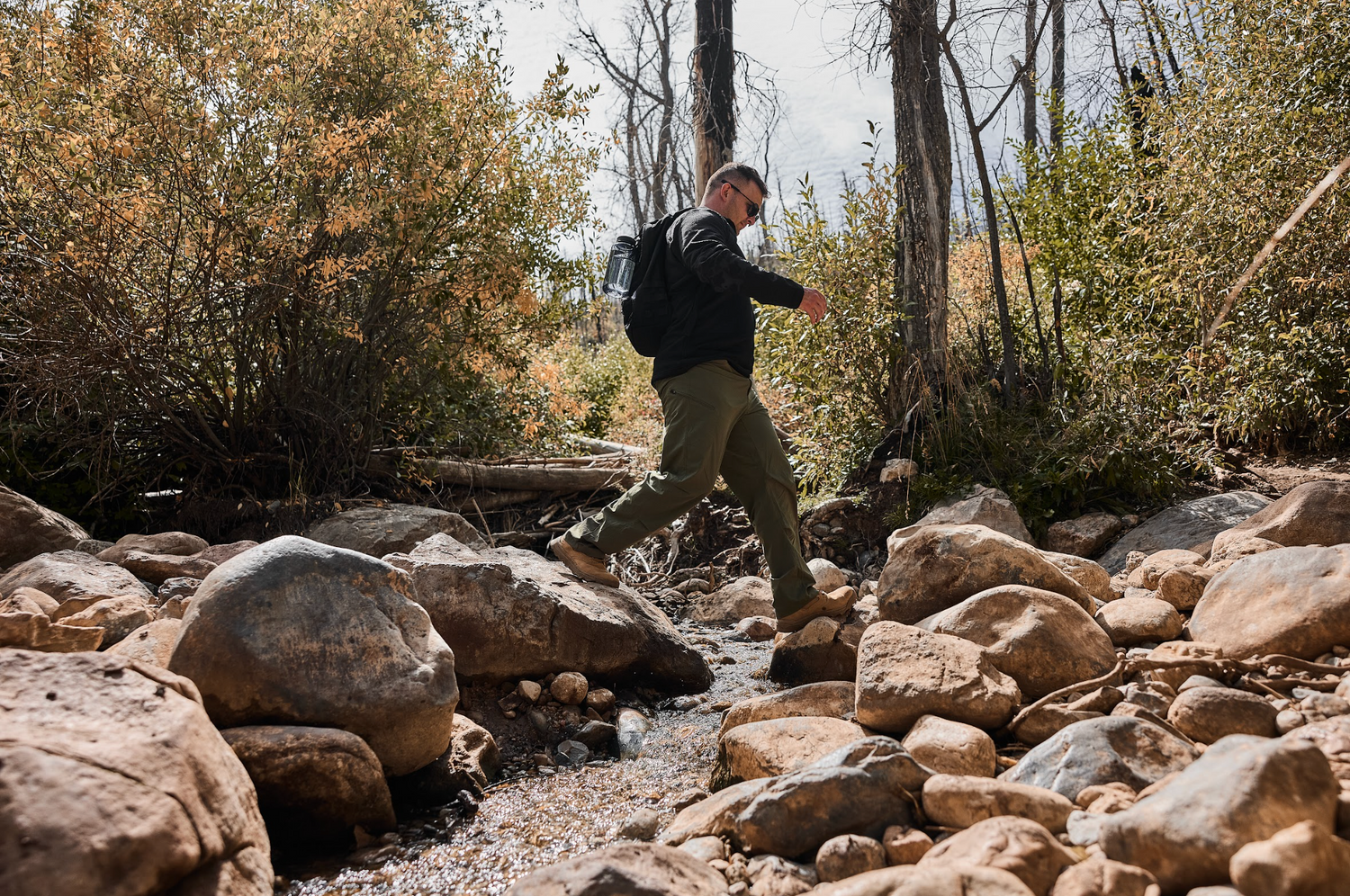 Man rucking with GORUCK backpack across rocky stream in outdoor wilderness setting