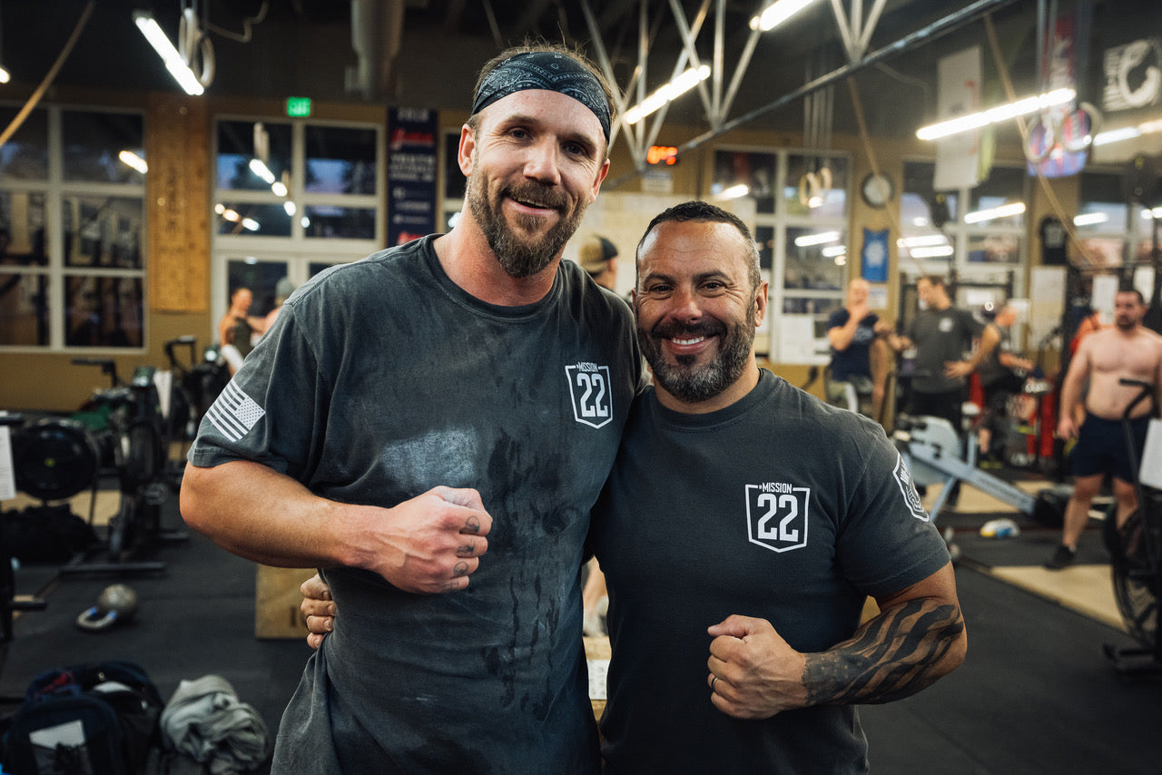 Two men wearing Mission 22 shirts smiling in a GORUCK gym, post-workout, with gym equipment and people in the background.