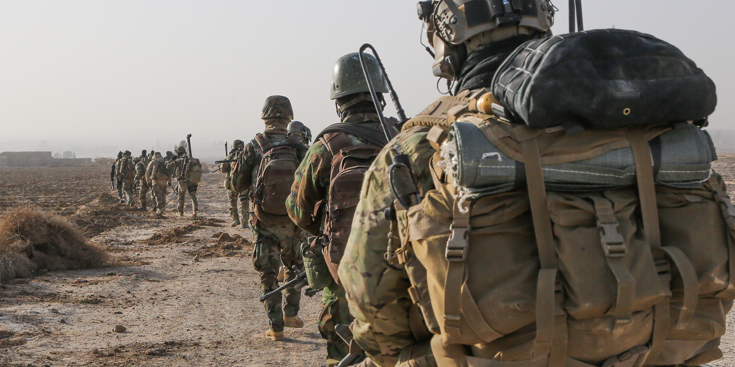 Group of soldiers in camouflage gear rucking with backpacks on a dirt trail, GORUCK event