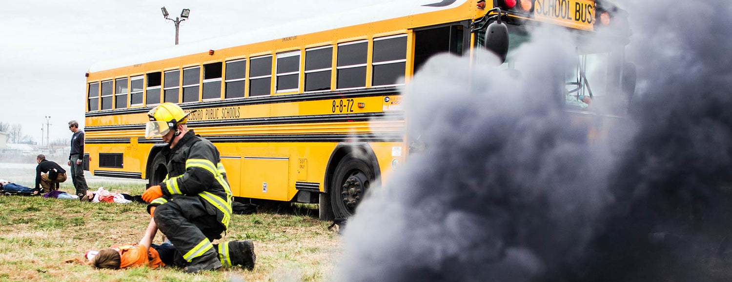 Firefighter assists injured person near yellow school bus with smoke, emergency training scene