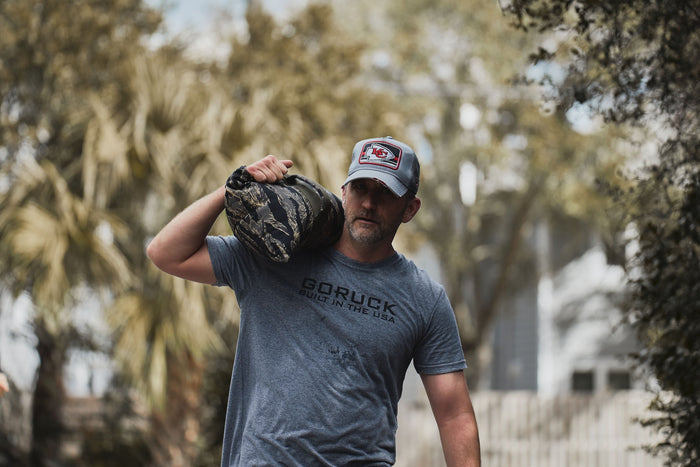Man outdoors training with GORUCK sandbag, wearing GORUCK shirt and cap, promoting rucking gear