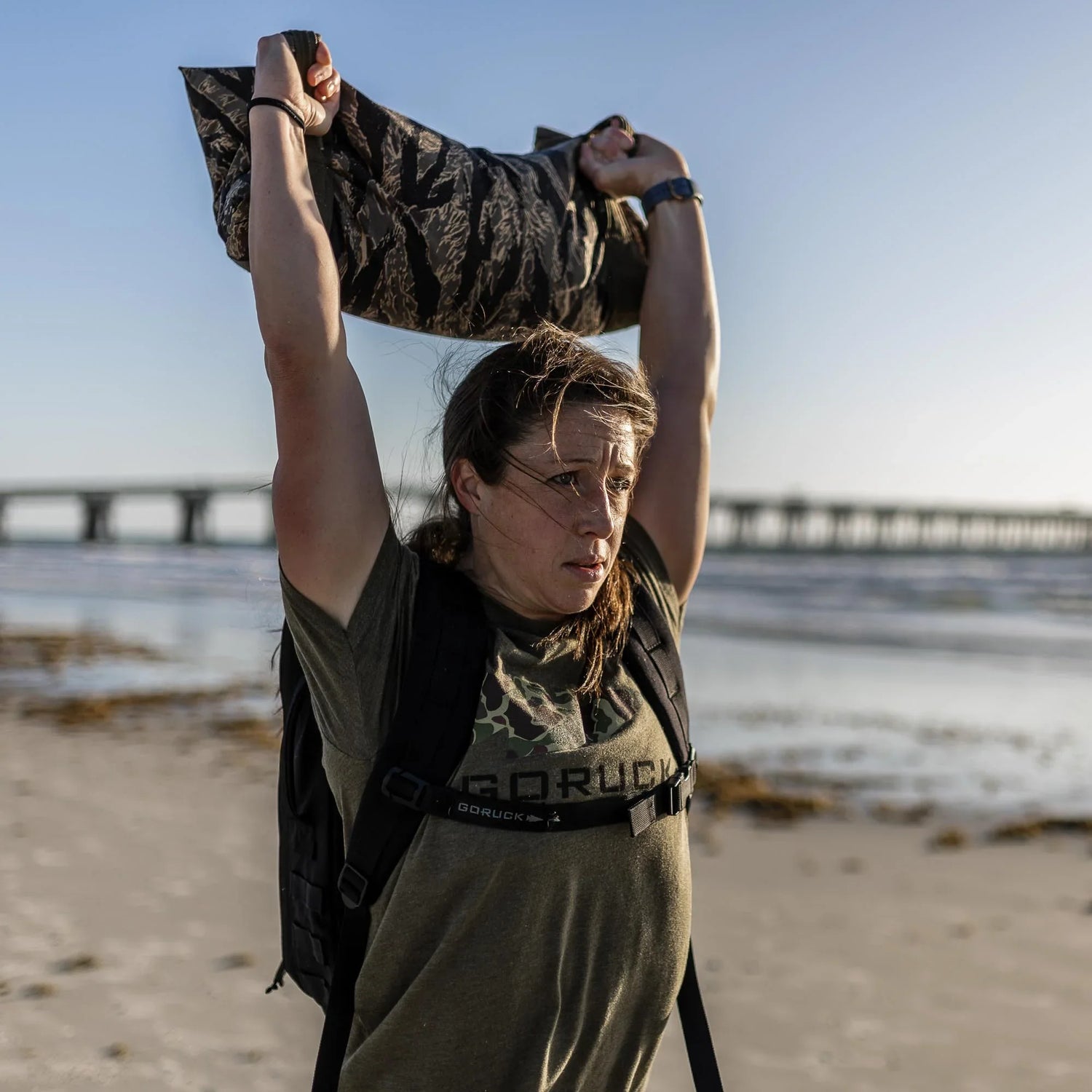 Woman on beach lifting weighted sandbag overhead wearing GORUCK gear, bridge in background