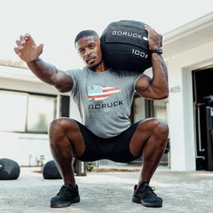 Man squatting outdoors with GORUCK 100 lb sandbag on shoulder, wearing GORUCK shirt