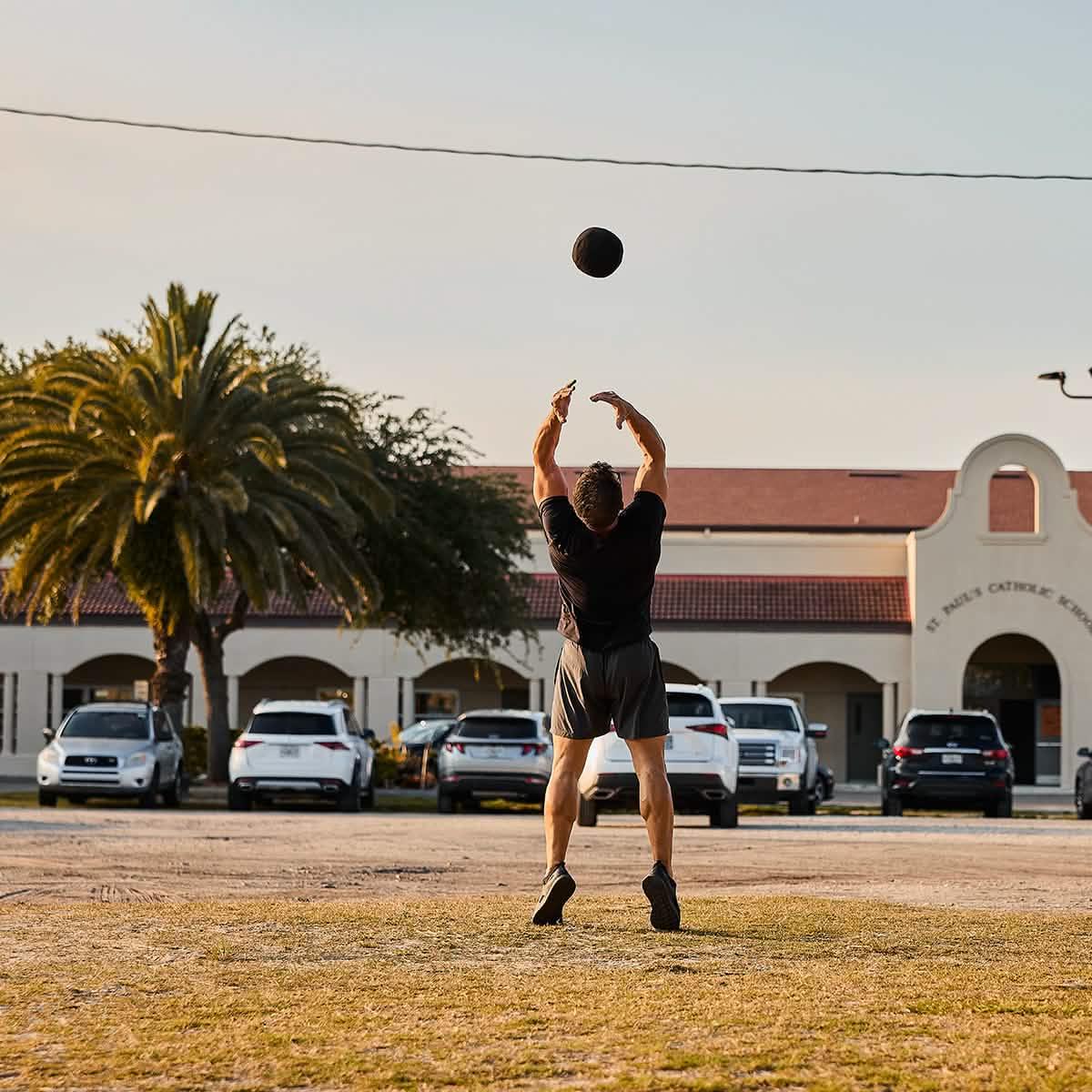 Man outdoors throwing weighted ball, GORUCK rucking workout with school and cars in background
