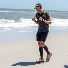 A man jogs on a sandy beach near the ocean, sporting a backpack, cap, sunglasses, athletic gear, and Merino Compression Socks for maximum comfort.