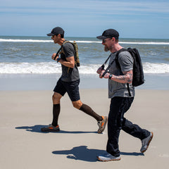 Two men jog along a sandy beach near the ocean under a clear sky, both wearing Merino Compression Socks designed for graduated compression.