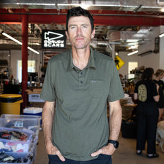 A man in an olive green Men's Polo with Pocket - Tri-BlendX stands in a busy workspace with a “GORUCK SCARS” sign in the background, highlighting the polo’s comfort and anti-stink features.