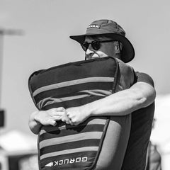 Man carrying GORUCK sandbag outdoors, wearing sunglasses and a tactical hat