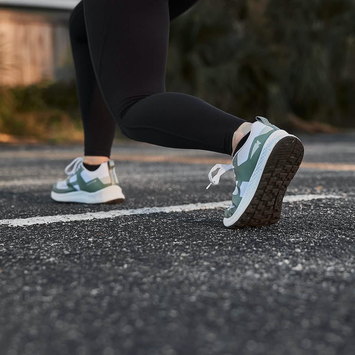 A woman in black leggings, wearing a pair of GORUCK Women's Rough Runner - Seaspray sneakers featuring a Gradient Density EVA Midsole, is stretching or getting ready to run on an outdoor paved surface. The focus is on her shoes as sunlight casts a warm glow over the scene.