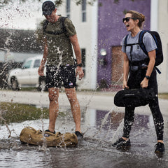Two people with backpacks laugh as water splashes around them in a puddle on a suburban street, both wearing Men’s USA Training Shorts - Flag - ToughStretch from GORUCK.