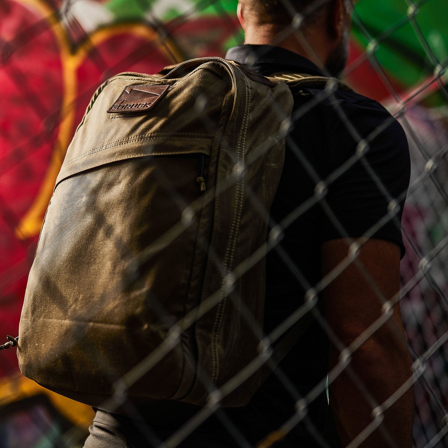 A man with the GR1 USA - Heritage Waxed Canvas rucksack stands behind a wire fence, with colorful graffiti blurred in the background.