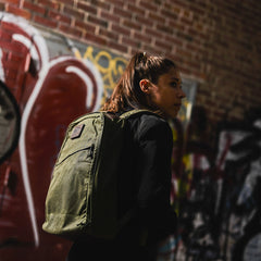 A woman with a ponytail stands by graffiti-covered brick walls in dim lighting, carrying the GR1 USA - Heritage Waxed Canvas backpack.