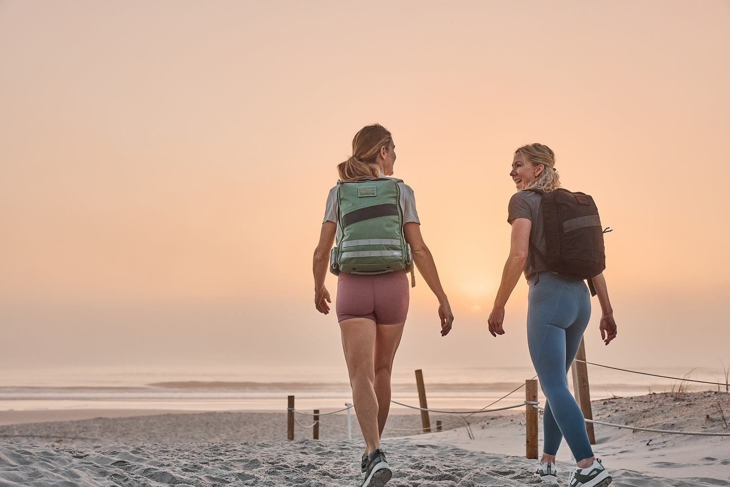Two women with backpacks walk on a sandy beach at sunset, smiling and talking together.