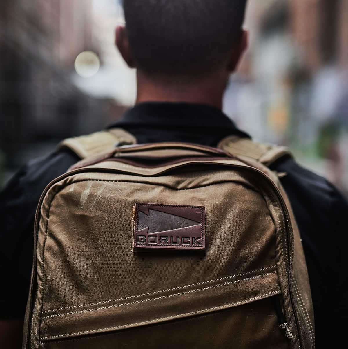Person wearing a GORUCK tan backpack in an urban outdoor setting, close-up view