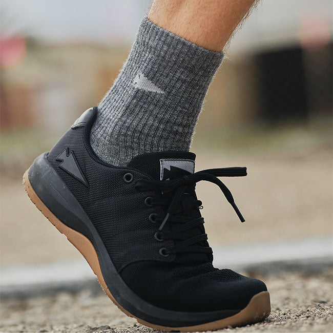 Close-up of a person wearing gray socks and black athletic shoes with brown soles, walking outdoors.