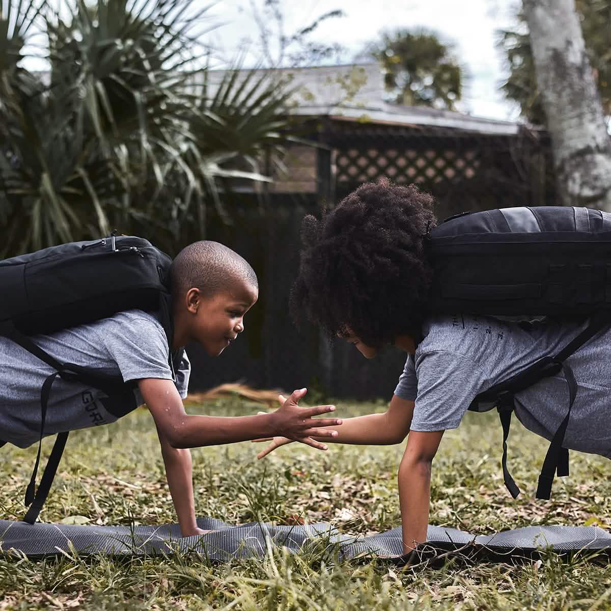 Two kids wearing black GORUCK backpacks do push-ups and high-five outdoors on grass.