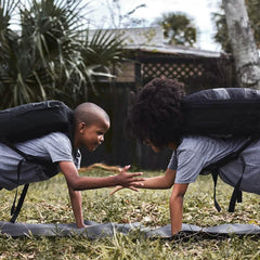 Two kids wearing black GORUCK backpacks do push-ups and high-five outdoors on grass.