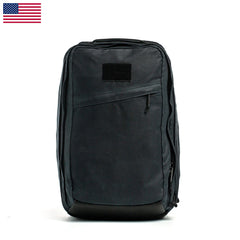 The GR2 - Heritage Waxed Canvas backpack in black features a front zipper pocket and a US flag at the top left, inspired by rugged travel rucks, displayed on a white background.