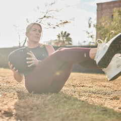 Woman outdoors using GORUCK exercise gear, performing core workout on grass in sunlight