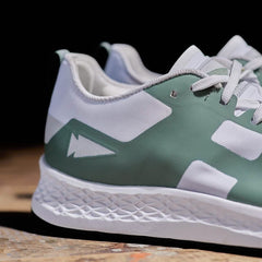 A close-up of the Women's Rough Runner - Seaspray sneaker by GORUCK highlights its green and white design. The shoe is adorned with a stylized lightning bolt symbol on the side and features a textured white sole equipped with a Gradient Density EVA Midsole. The dark background accentuates the intricate details of the shoe, making it stand out.