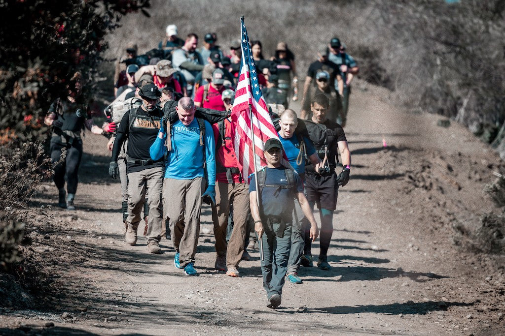 A group of hikers, one carrying an American flag, walk together on a dirt trail through rugged terrain.