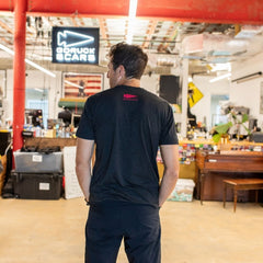 A man stands in an office, facing away, wearing a black Buddy FCKR Friday t-shirt beneath a neon GORUCK SCARS sign.