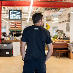 A man in a black “Easy Life Is Not For Us” t-shirt stands in an industrial-style room with a GORUCK SCARS sign and an American flag on the wall.
