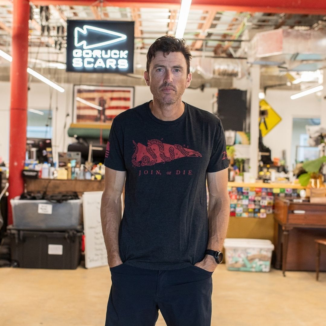 A man stands in a workshop, hands in his pockets and looking at the camera, wearing the Join or Die Tee - Tri-Blend inspired by Ben Franklin’s iconic “Join or Die” cartoon—a timeless emblem of colonial unity.