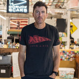 A man stands in a workshop, hands in his pockets and looking at the camera, wearing the Join or Die Tee - Tri-Blend inspired by Ben Franklin’s iconic “Join or Die” cartoon—a timeless emblem of colonial unity.