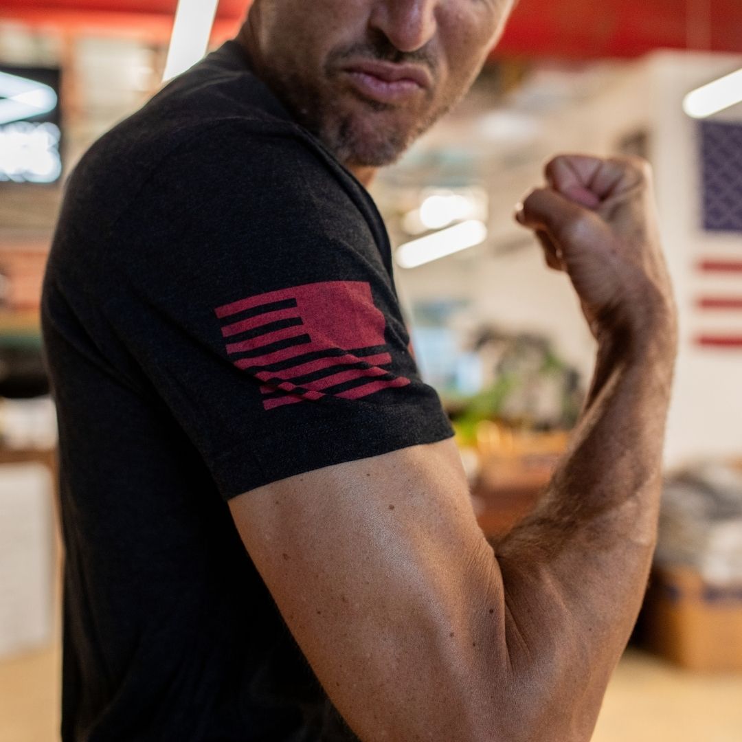 A man flexes his arm indoors, wearing the Join or Die Tee - Tri-Blend, a black shirt featuring a red flag design inspired by Ben Franklin’s iconic cartoon, with a blurred background.