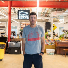 A man wearing the “100% American” Graphic T-Shirt stands in an industrial office space with colorful decor and an exposed ceiling.