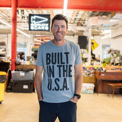 A man smiles indoors while wearing the Graphic T-Shirt - Built in the USA (gray) in a modern workspace.