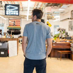 A man in a gray Join or Die Tee - Tri-Blend stands in a workshop, facing away, surrounded by equipment and a “GORUCK SCARS” sign—an atmosphere that echoes Ben Franklin’s era of craftsmanship and colonial unity, channeling the spirit of the legendary “Join or Die” cartoon.