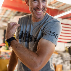 A smiling man flexes his arm while wearing the Join or Die Tee - Tri-Blend in gray, featuring “250” on the sleeve and paired with a yellow watch, evoking Ben Franklin’s iconic Join or Die cartoon symbolizing colonial unity.