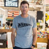 A man in a gray “Join or Die Tee - Tri-Blend,” featuring the iconic Ben Franklin cartoon symbolizing colonial unity, stands in a casual workspace with a “GORUCK SCARS” sign.