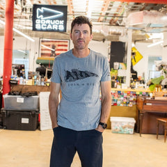 A man in a gray “Join or Die Tee - Tri-Blend,” featuring the iconic Ben Franklin cartoon symbolizing colonial unity, stands in a casual workspace with a “GORUCK SCARS” sign.